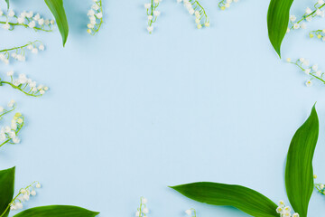 small white lilies of the valley with large green leaves lie on a pastel blue background with empty spaces in the center. Top view, concept for spring holidays, template blank