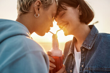 Happy young lesbian couple having romantic moment, drinking from one glass bottle with the straw,...