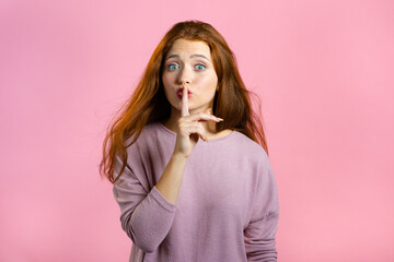 Smiling woman with red hair holding finger on her lips over pink background. Gesture of shhh, secret, silence. Close up.