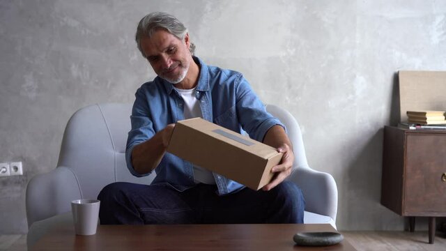 Smiling Grey-haired Mature man customer opening parcel cardboard box sitting at home. Happy consumer unpacking postal shipping delivery satisfied with good purchase. Fast post shipment service concept