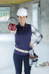 smiling female construction worker wearing helmet shows protective headphones