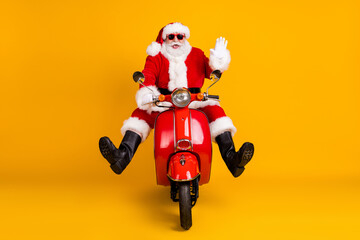 Portrait of his he nice funny comic childish cheerful cheery Santa riding motor bike traveling having fun waving hello grimacing fast speed isolated bright vivid shine vibrant yellow color background
