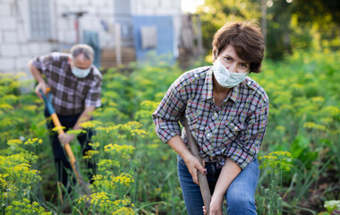 Female farmer in protective mask with shovel in the backyard of country house