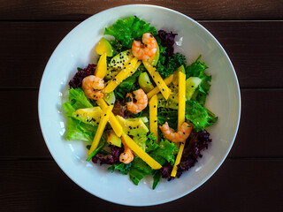 appetizing salad of shrimp, mango, avocado, cucumber, mix of lettuce leaves, decorated with black sesame seeds, on a white plate, flatlay