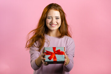 Excited woman holding gift box and gives it by hands to camera on pink wall background. Girl smiling, she is happy with present. Studio portrait