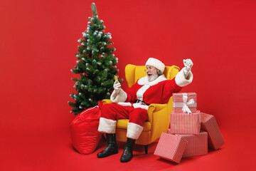 Excited Santa Claus man in Christmas hat suit sit in armchair with fir tree gifts using mobile phone doing winner gesture isolated on red background. Happy New Year celebration merry holiday concept.