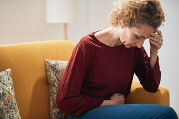 Woman with stomach pain and headache problem sitting on a couch at home.