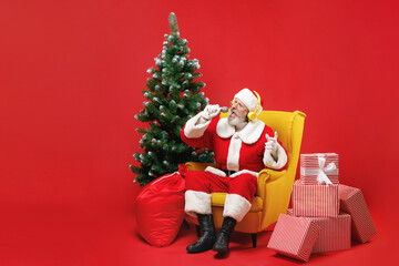 Fototapeta na wymiar Funny Santa Claus man in Christmas suit sit in armchair with fir tree gifts listen music with headphones sing song on microphone isolated on red background. Happy New Year celebration holiday concept.
