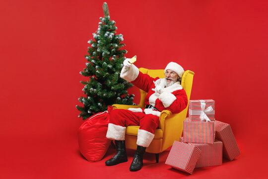 Excited Santa Claus man in Christmas hat suit sit in armchair with fir tree gifts doing selfie shot pointing on mobile phone isolated on red background. Happy New Year celebration holiday concept.