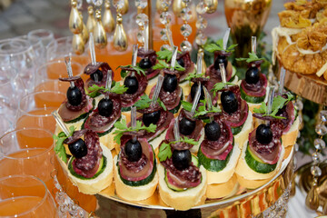 Sandwiches on the buffet table. Small canapes with sausage, cheese and olive. Sandwiches with sausage and olive are on a golden platter, surrounded by glasses of juice.