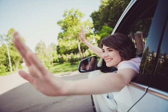 Photo of cheerful attractive smiling young girl lady look out car window wave hi two hands people fans celebrity show careless trick trend move make photo new auto wear pink t-shirt green area outside
