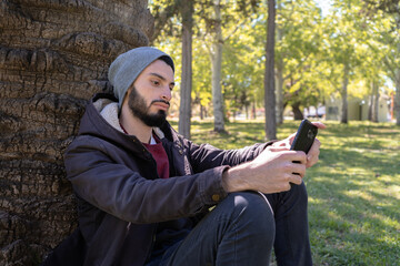 Man outdoors sitting on the ground against a tree using his smartphone