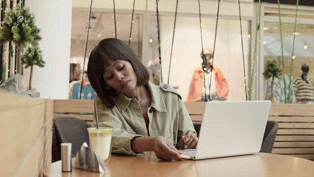 Medium shot of middle-aged African woman wearing casual clothes sitting at cafe table in mall in front of laptop, inserting flash drive into computer and starting typing on keyboard