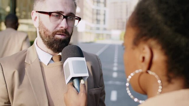 Over the shoulder shot of Caucasian businessman standing outdoors in the city and talking in microphone while giving TV interview to Afro-American female journalist