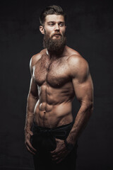 Fototapeta na wymiar Brutal bearded guy with naked torso and muscular build with hands in pockets posing in dark studio background.