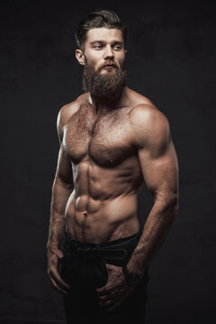 Brutal bearded guy with naked torso and muscular build with hands in pockets posing in dark studio background.