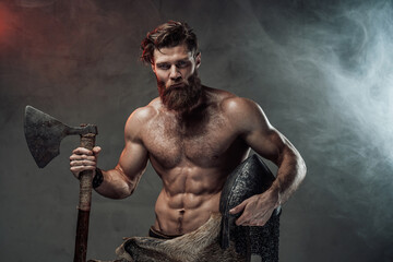 Serious and muscular viking with beard and naked torso staying in dark background with lights and...
