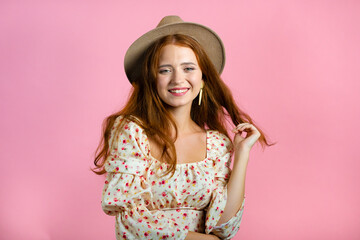 Young woman with long red wavy and shiny hairstyle and hat looking to camera, flirting. Beautiful model girl demonstrate her elegant volume hair. Pink studio background.