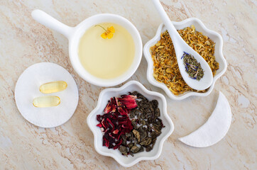 Small white bowls with cosmetic (massage, cleansing) aroma oil, dry marigold flowers, healing herbs, vitamin E capsules, green tea, hibiscus, cotton pad, eye patch. Natural skin care, homemade spa, be
