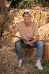 Naklejka na ściany i meble Male farmer sitting on hay, holding clip folder and bunch of millet spikelets