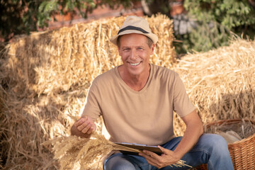Naklejka na ściany i meble Male farmer sitting on hay, holding clip folder and bunch of millet spikelets, smiling