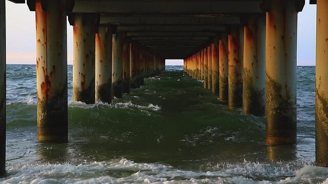 Rushing sea under the pier columns in the evening at sunset. Waves crashing against the old rusty piers of the pier