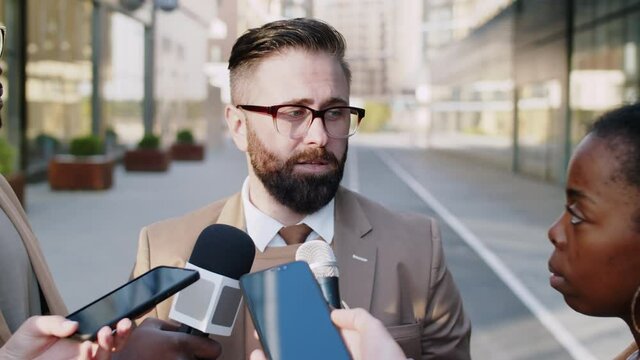 Chest up shot of Caucasian businessman standing outdoors in the city surrounded by multiethnic journalists holding smartphones and microphones, looking at camera and speaking while giving interview