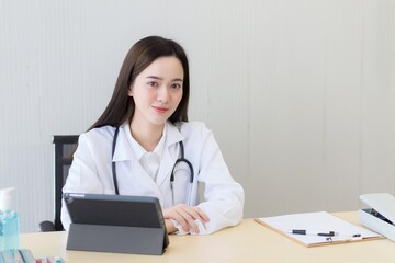 Asian beautiful young smiling female doctor sitting in office at hospital. On table has a clipboard and digital tablet computer.