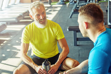 Positive middle aged man in sportswear holding bottle of water, talking with his personal trainer or fitness instructor and smiling while exercising at gym or sport club