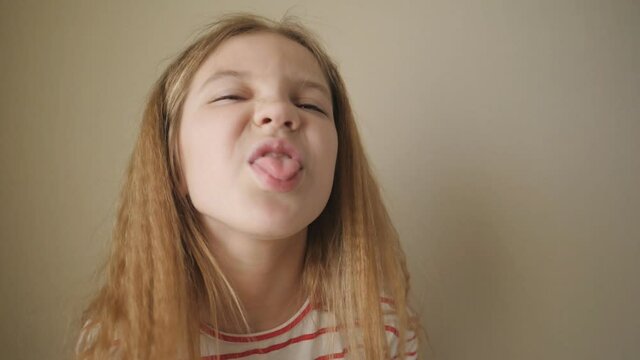 Small female child sticking her tongue out and turning aside indoor. Little girl with long blonde hair showing displeased emotion into camera. Cute kid fooling around against the background of wall