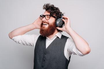 Bearded cheerful young man is listening to the music with headphones on holding them with both of his hands near a white wall