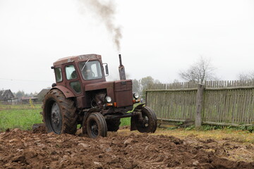 Old Soviet AWD wheeled brown diesel tractor with exhaust smoke plowing the field on backyard close up, soil cultivation on an autumn day on woden fence background, rural agriculture farming landscape