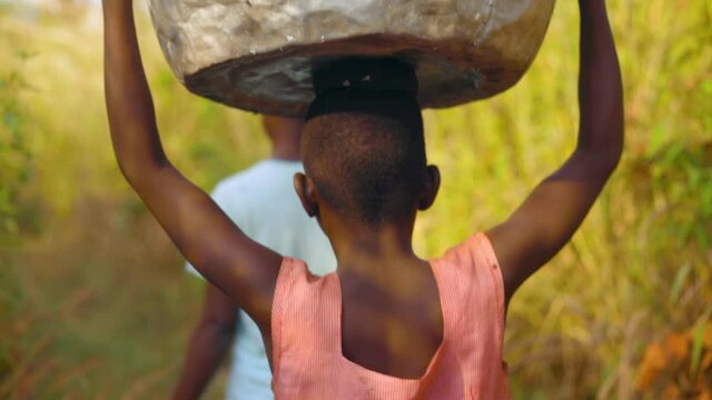 Group of local young African women and kids are carrying big heavy buckets of drinking water and bringing the water on their heads towards the village walking in bush field - close shot