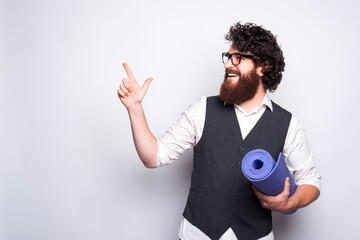 Young bearded man is holding a yoga matt and pointing at a free space is smiling near a white wall