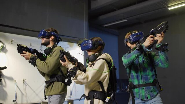 Medium shot of team of three young friends, two men and women, in virtual reality headsets going into attack with gun controllers while playing VR adventure game
