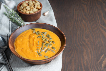 Vegetarian pumpkin soup made of pureed vegetables cooked on thanksgiving decorated with seeds and thyme served in bowl on dark wooden background with spoon, textile and croutons. Image with copy space