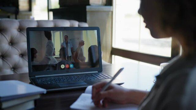 online webinar, young female student undergoes remote training via video communication on laptop raises hand and answers question by making notes in notebook sitting at table by window