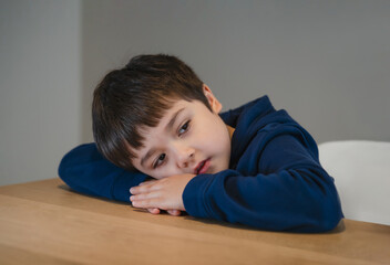 Portrait tired kid lying head down on his arm lookig out deep in throught, Bored child laying head on table with unhappy face, Lonely Young boy with sad face sitting alone, Health metal concept