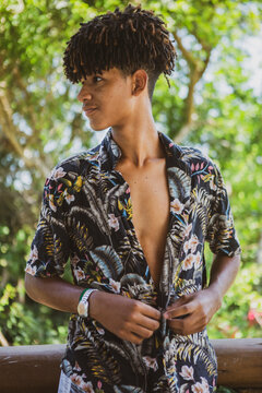Young African Brazilian man with trendy hairstyle unbuttoning his shirt and looking to the side in front of an out of focus vegetation background. New normal isolation concept.