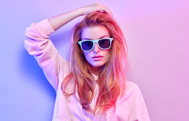 Fashionable hipster woman in Trendy outfit, stylish hair, makeup on pink purple neon light. Redhead model in hoodie, fashion sunglasses. Beautiful girl in autumn fall neon style