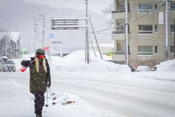 Picture of ski resort town while snowing and the street is covered with snow and a woman wear dark green jacket carring ski walking on the sidewalk.