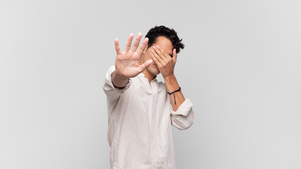 young handsome man covering face with hand and putting other hand up front to stop camera, refusing...