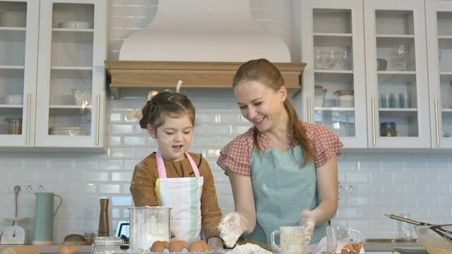 Cheerful little girl and young mommy throw flour making dough for tasty pie at large wooden table in contemporary kitchen