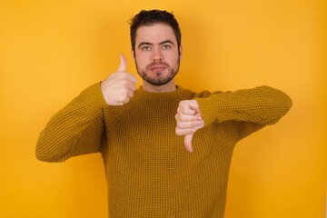 Young man wearing casual sweater and over isolated yellow background showing thumbs up and thumbs down, difficult choose concept - obrazy, fototapety, plakaty