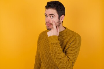 Young man wearing casual sweater and over isolated yellow background Pointing to the eye watching you gesture, suspicious expression.