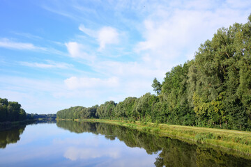 The Danube flows calmly and broadly through Bavaria, in front of green trees and under a blue sky with clouds