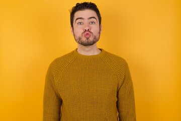 Shot of pleasant looking Young man wearing casual sweater and over isolated yellow background, pouts lips, looks at camera, Human facial expressions