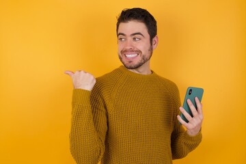Portrait of Young man wearing casual sweater and over isolated yellow background using and texting with smartphone  pointing and showing with thumb up to the side with happy face smiling