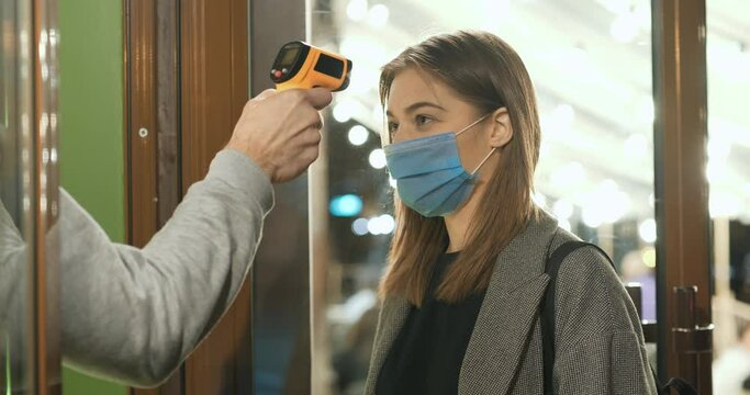 Slow Motion Taking body temperature before entering a restaurant. Screening of people for Covid-19 coronavirus symptoms. Young woman in medical protective mask enters the store