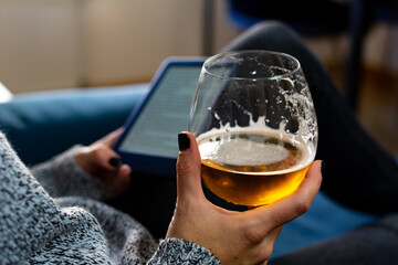 A woman with painted nails, holding a glass of beer in the foreground, can be seen in the background holding an e-book with the other hand. All of this while she rests on an armchair at home